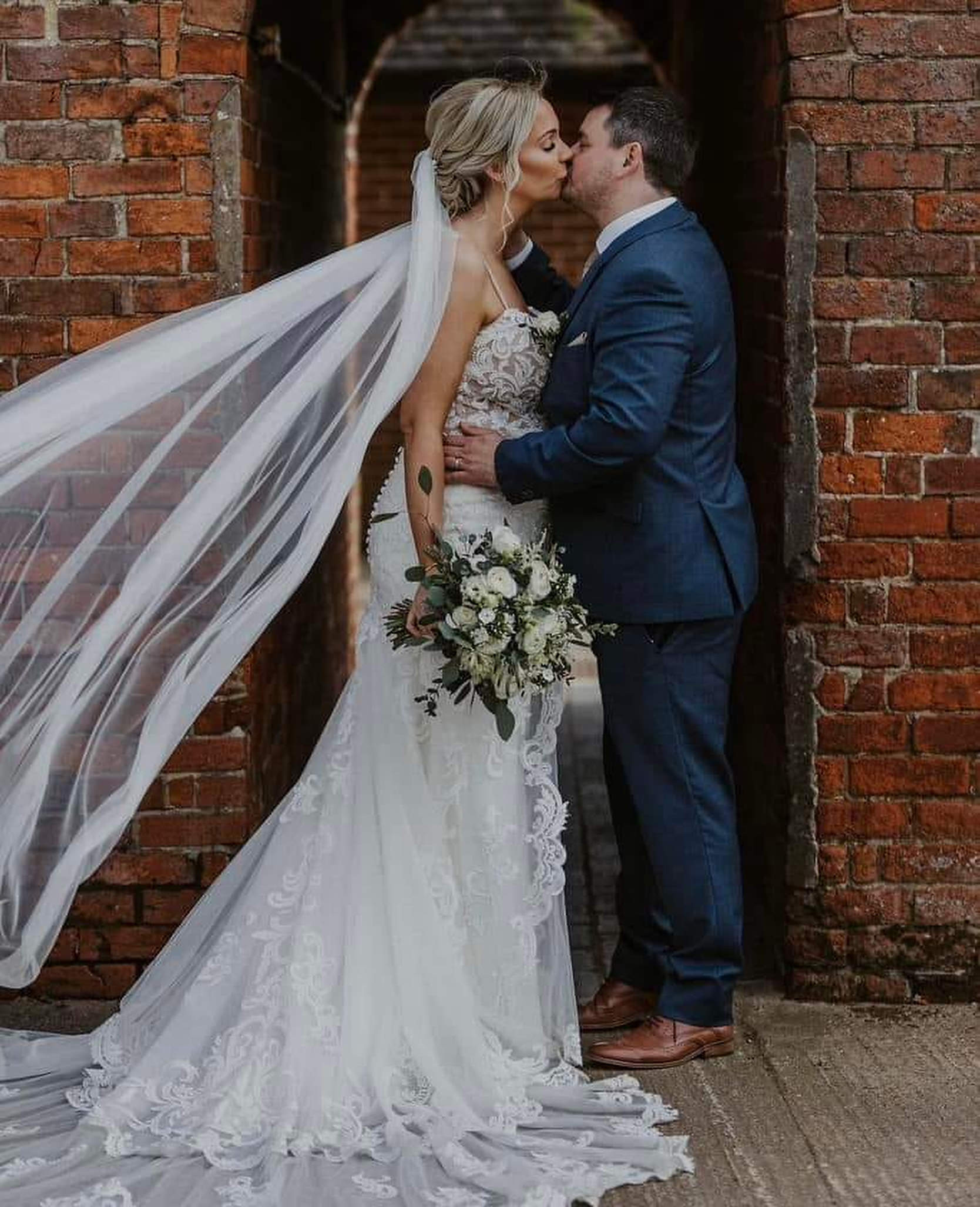 Portrait of the newly married couple kissing with a flowing veil and a stunning bouquet of flowers in hand, complimented perfectly by the buttonhole on the suit